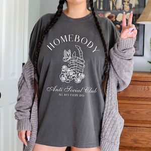 May include: A dark gray t-shirt with a white graphic design. The graphic features a skeleton hand holding a butterfly with flowers around it. The text "Homebody Anti Social Club All Day Every Day" is printed on the shirt.