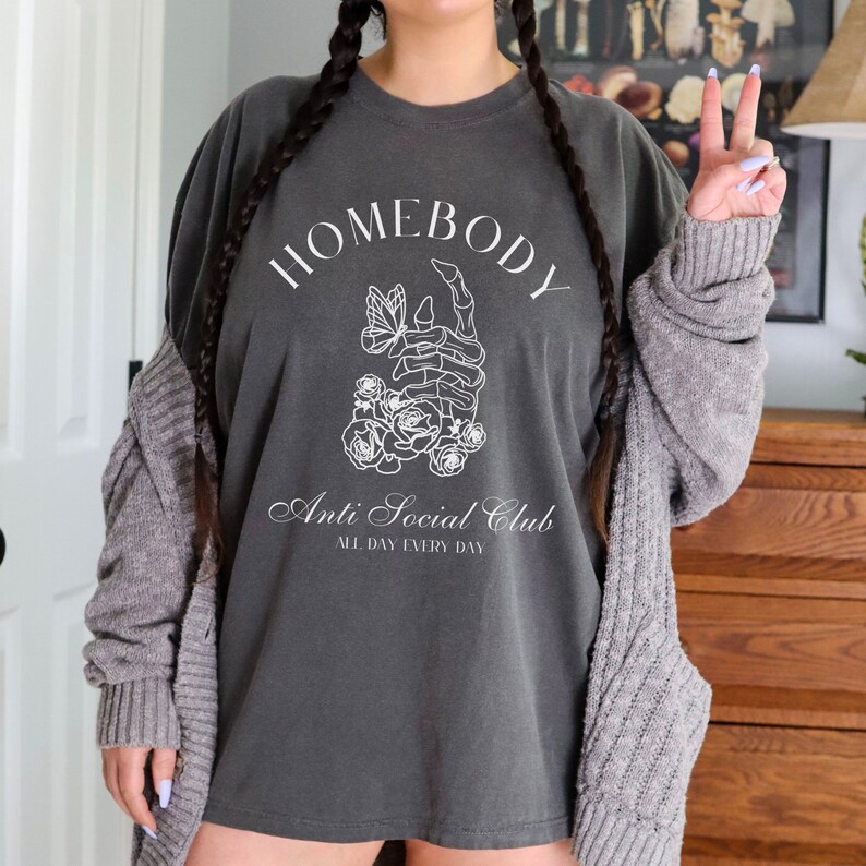 May include: A dark gray t-shirt with a white graphic design. The graphic features a skeleton hand holding a butterfly with flowers around it. The text "Homebody Anti Social Club All Day Every Day" is printed on the shirt.
