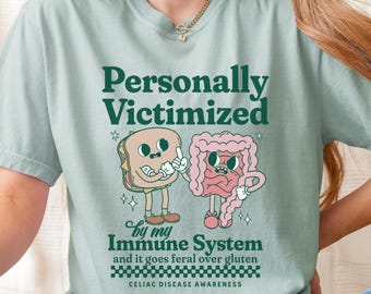 May include: A light blue t-shirt with a graphic of a sandwich and a cartoon intestine. The text on the shirt reads "Personally Victimized by my Immune System and it goes feral over gluten. Celiac Disease Awareness."