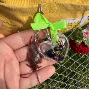 May include: A keychain with a clear heart-shaped charm containing a black spider and a black 8-ball. A brown cockroach charm and a green bow are also attached. The keychain is attached to a silver clasp.