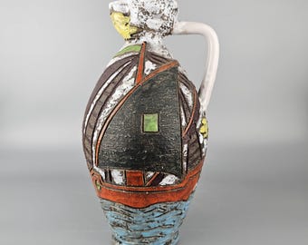 Puede incluir: Jarra de cerámica con diseño de barco. La jarra tiene un asa blanca y una superficie texturizada. El diseño del barco incluye una vela negra, detalles en naranja y agua azul en la base. La parte superior de la jarra tiene un diseño floral blanco y amarillo.