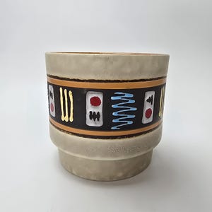 Op de afbeelding: Een cilindervormige keramische plantenpot met een getextureerde beige afwerking. Een brede zwarte band omcirkelt het midden, met een herhalend patroon van witte rechthoeken met rode cirkels, gele verticale lijnen en lichtblauwe zigzaglijnen.