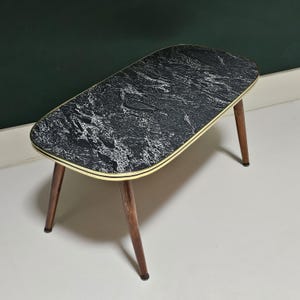 Puede incluir: Una mesa de centro de mediados de siglo con una tapa negra y blanca con efecto mármol y un borde dorado. La mesa tiene cuatro patas de madera. La mesa mide aproximadamente 76 cm de largo, 38 cm de ancho y 46 cm de alto.
