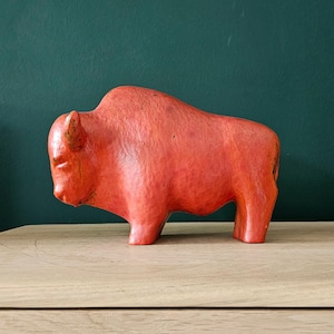Può includere: Una statuetta di bisonte color corallo con una superficie strutturata. La scultura è appoggiata su una superficie di legno colorata, con uno sfondo verde scuro. Il bisonte è un oggetto decorativo.