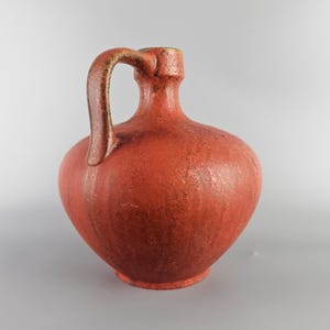 Puede incluir: Una jarra de cerámica roja con cuerpo redondeado y asa curva. La jarra tiene una superficie texturizada, ligeramente moteada.