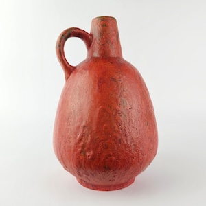 Puede incluir: Jarra de cerámica roja texturizada con asa. La jarra tiene un cuerpo bulboso y un cuello estrecho. El asa es arqueada y está unida al lateral. La jarra es de color rojo sólido con algunas zonas más oscuras.