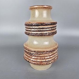 Puede incluir: Un jarrón de cerámica beige con un diseño texturizado y acanalado en tonos marrones. El jarrón tiene una forma redondeada con un cuello estrecho y una base ancha. Las bandas texturizadas rodean el jarrón, creando un atractivo visual único. El jarrón mide unos 25 cm de alto.