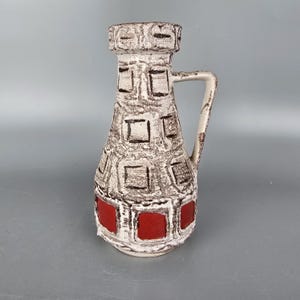 Puede incluir: Un jarrón de cerámica vintage con asa, con una base crema con patrones geométricos marrones y rojos. El jarrón tiene forma cilíndrica con superficie texturizada y parte superior decorativa.