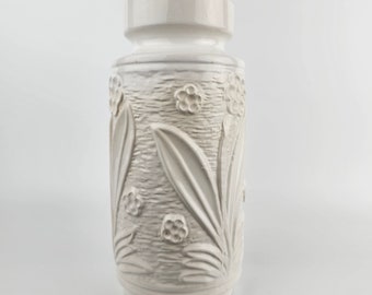 May include: White ceramic vase with an embossed floral design. The vase has a textured surface and a cylindrical shape.