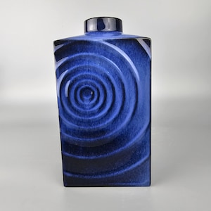 Könnte beinhalten: Eine rechteckige, kobaltblaue Keramikvase mit glänzendem Finish. Die Vase hat ein Spiraldesign auf der Vorderseite, mit einer dunkelblauen, rechteckigen Oberseite und einem dunkelblauen, zylindrischen Verschluss.