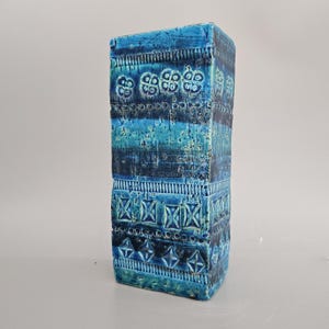 May include: A tall, rectangular blue ceramic vase with a geometric pattern. The vase has a textured surface and is decorated with a variety of blue and green shades.