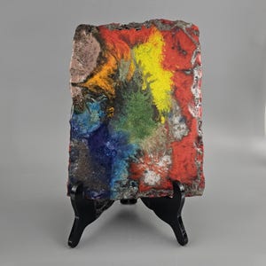 Puede incluir: Una pintura abstracta colorida sobre un lienzo texturizado de forma irregular. La pintura presenta salpicaduras de pintura roja, amarilla, verde, azul y blanca. El lienzo se muestra en un caballete negro.