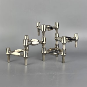 Può includere: Tre candelabri in metallo argentato. Ogni portacandela ha un design scultoreo unico con forme curve e geometriche. I portacandele sono disposti su uno sfondo grigio neutro, mettendo in risalto i loro dettagli intricati e le superfici riflettenti.