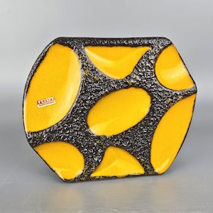 Puede incluir: Un plato hexagonal de cerámica con un fondo negro texturizado y acentos amarillos brillantes de forma irregular. El plato tiene un acabado brillante y una pequeña etiqueta que dice "rott keramik".