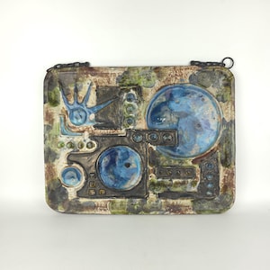 May include: A rectangular ceramic wall plaque featuring an abstract relief design with a mottled brown, green, and cream background. The surface displays raised geometric shapes, circular indentations, and a prominent blue hand-like motif.