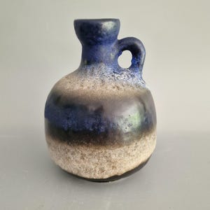 May include: A ceramic vase with a rounded body and a handle. The vase features a gradient of colors, including blue, brown, and beige. The top and handle are a deep blue, transitioning to brown and beige at the base. The vase has a textured surface.