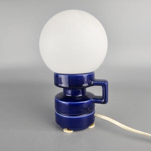 Puede incluir: Lámpara de mesa vintage con una pantalla esférica de vidrio blanco y una base de cerámica azul oscuro. La base tiene forma cilíndrica con un asa y pequeños pies blanquecinos. La lámpara está enchufada.