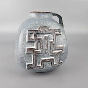 May include: A light blue ceramic vase with a handle and a textured surface. The vase features a raised, geometric design in shades of brown and white. The vase has a rounded body and a small opening.