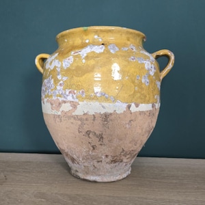 Puede incluir: Una gran olla de cerámica amarilla y marrón antigua con dos asas. La olla tiene un aspecto envejecido y desgastado con pintura desconchada y una franja blanca en el medio. La olla probablemente se utiliza con fines decorativos.