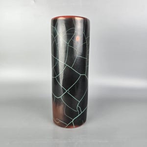 Könnte beinhalten: Eine hohe, zylindrische Vase mit einem Craquelé-Glasurmuster. Die Vase hat eine dunkle, glänzende Oberfläche mit einem Netzwerk aus türkisfarbenen Linien. Der obere Rand ist rotbraun, und die Basis hat einen ähnlichen Farbton. Ein dekorativer Artikel.