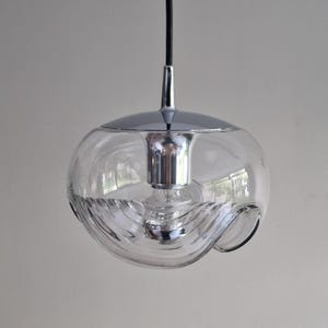 May include: A clear glass pendant light with a chrome finish. The light has a spherical shape with a textured, wave-like design on the lower half. A black cord suspends the light from the ceiling.