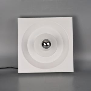 Puede incluir: Una lámpara de techo cuadrada blanca con un diseño de círculos concéntricos. El centro presenta una luz esférica cromada. La lámpara está montada sobre un fondo gris.