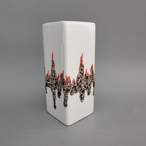 May include: White ceramic vase with a black and orange geometric design. The design features a series of jagged lines with a textured surface.