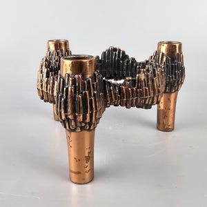 Può includere: Un candelabro in rame a tre braccia con un design testurizzato e geometrico. Ogni braccio ha un'apertura circolare per una candela.