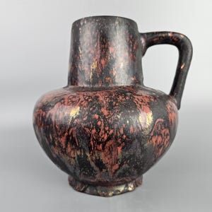 Könnte beinhalten: Eine schwarze Keramikkanne mit einer rot-goldenen Marmorierung. Die Kanne hat einen runden Körper und einen einzigen Henkel.