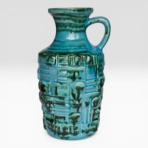 Può includere: Un vaso in ceramica turchese con manico. Il vaso ha una superficie strutturata con forme geometriche e organiche nelle tonalità del verde acqua e verde scuro. Il vaso è alto circa 25 cm.