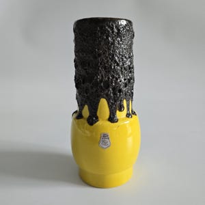 May include: A cylindrical ceramic vase with a bright yellow smooth base and a textured, dark brown volcanic glaze dripping from the top rim. A small white shield-shaped sticker with the text ES CERAMIA is centered on the yellow section.