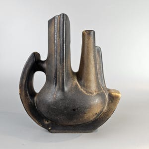 May include: A black and brown ceramic vase with an abstract, sculptural design. The vase has three openings, one with a handle, and a unique, asymmetrical shape.
