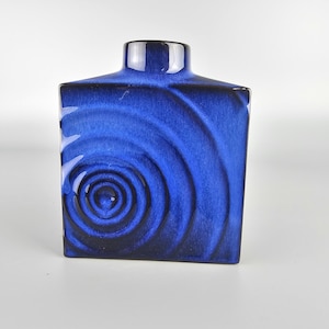 Könnte beinhalten: Eine quadratische, kobaltblaue Keramikvase mit glänzendem Finish. Die Vase hat ein Spiraldesign auf der Vorderseite, das einer stilisierten Rose ähnelt. Die Oberseite der Vase hat eine rechteckige Öffnung und einen dunkelblauen Rand.