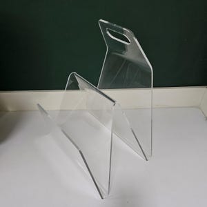 May include: A clear acrylic magazine rack with two tiers and a handle on the top. The rack is designed to hold magazines, newspapers, or other reading materials.