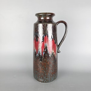 May include: A tall, ceramic vase with a handle. The vase has a speckled brown and teal base, with vertical red and white streaks. The rim and handle are a darker brown. The vase is approximately 30 cm tall.