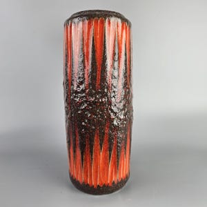 May include: A tall, cylindrical ceramic vase with a textured, brown and orange glaze. The vase has a geometric pattern of red and black triangles.