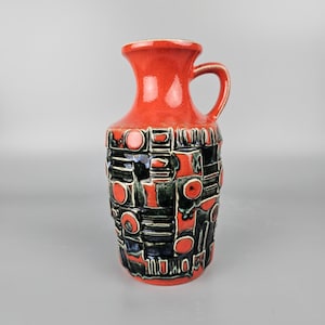Può includere: Un vaso in ceramica rosso con un motivo geometrico in nero e verde. Il vaso ha un manico e un collo stretto.