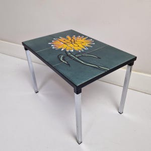 Peut inclure: Une petite table d'appoint carrée avec un cadre en métal chromé et un plateau en céramique. Le plateau en céramique présente un motif floral stylisé avec une fleur jaune et des feuilles vertes sur un fond bleu-vert.