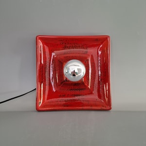 May include: A square, red wall sconce with a textured finish and a chrome-colored, spherical light fixture in the center. The sconce has a black power cord extending from the bottom left corner.