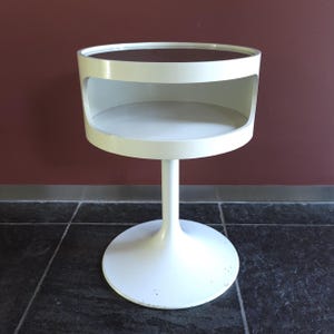 May include: A white circular side table featuring a pedestal base and a cylindrical upper section with an open storage compartment. The top surface is flat and circular, supported by a central column that connects to a wide, stable round base.