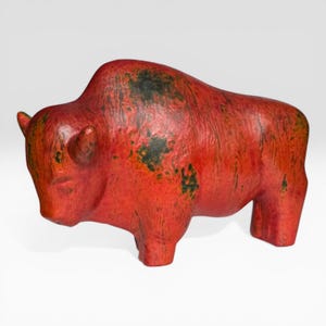 Può includere: Statuetta di bisonte rosso con macchie verde scuro e nere. La scultura ha una superficie strutturata e si trova su uno sfondo bianco. Il bisonte ha piccole corna e un corpo arrotondato.