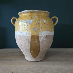 Könnte beinhalten: Ein großer, antiker Keramiktopf in Gelb und Beige mit zwei Henkeln. Der Topf hat ein abgenutztes, gealtertes Aussehen mit abblätternder Farbe und einem weißen Streifen. Er wird wahrscheinlich zur Aufbewahrung oder Dekoration verwendet. Höhe: ca. 30 cm.