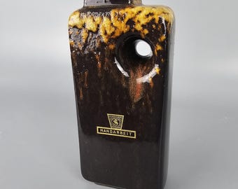May include: A rectangular brown ceramic vase with a large circular opening. The top and edges have a textured, golden-yellow glaze. The vase has a label that reads "HANDARBEIT".