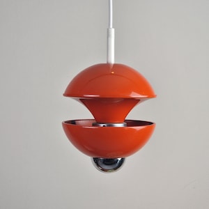 Op de afbeelding: Een retro-stijl oranje hanglamp met een witte kabel. De lamp heeft een gelaagd ontwerp met een afgeronde bovenkant, een conische middensectie en een komvormige bodem. Een chromen bol hangt eronder.