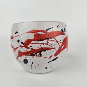 May include: White ceramic bowl with red and black splattered paint. The bowl has a rounded shape and a smooth, glossy finish.