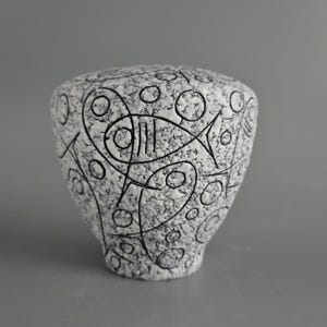 May include: A white ceramic vase with a textured surface and black abstract designs. The vase features a fish-like shape and other geometric patterns. The vase is a decorative item.