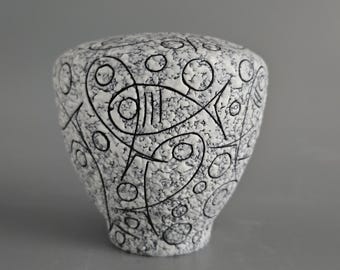 May include: A white ceramic vase with a textured surface and black abstract designs. The vase features a fish-like shape and other geometric patterns. The vase is a decorative item.