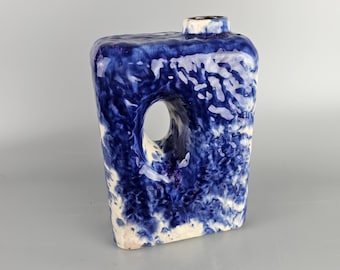 May include: A blue and white ceramic vase with a unique, abstract design. The vase has a hole cut out of the side and a textured surface.