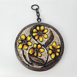 Puede incluir: Un colgante de pared redondo de cerámica con una cadena para colgar. La cerámica está decorada con un diseño floral estilizado en amarillo, naranja y verde.