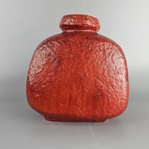 Puede incluir: Un jarrón de cerámica rojo con una superficie texturizada. El jarrón tiene una forma redondeada y bulbosa y una abertura ancha.